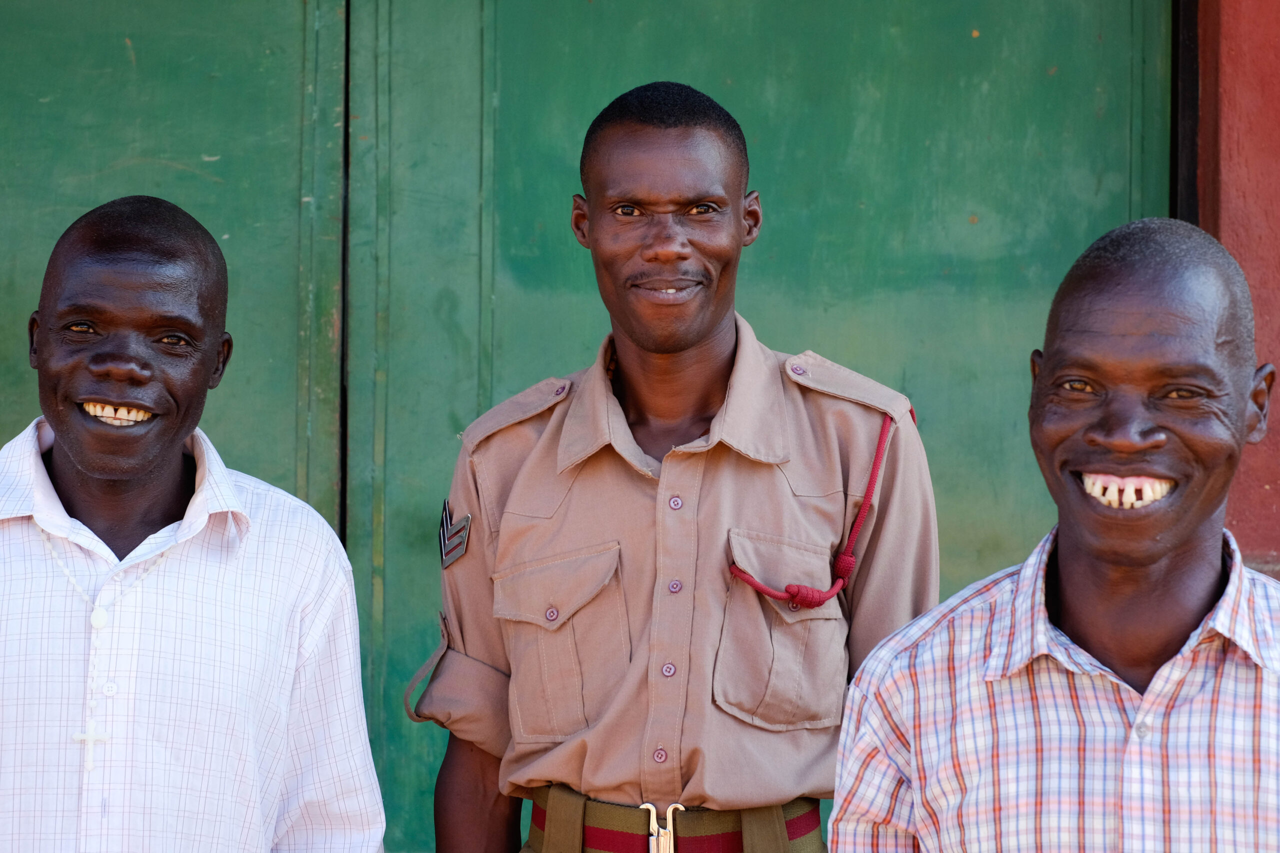 A man in a white shirt, a man in a beige uniform, and a man in a plaid shirt are looking at the camera and smiling in front of a green building.