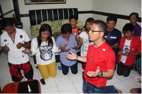 Nine people sit on their knees in a circle. Their eyes are closed and their heads tilt towards the ground. They are putting their arms in front of them and facing their hands towards the sky.